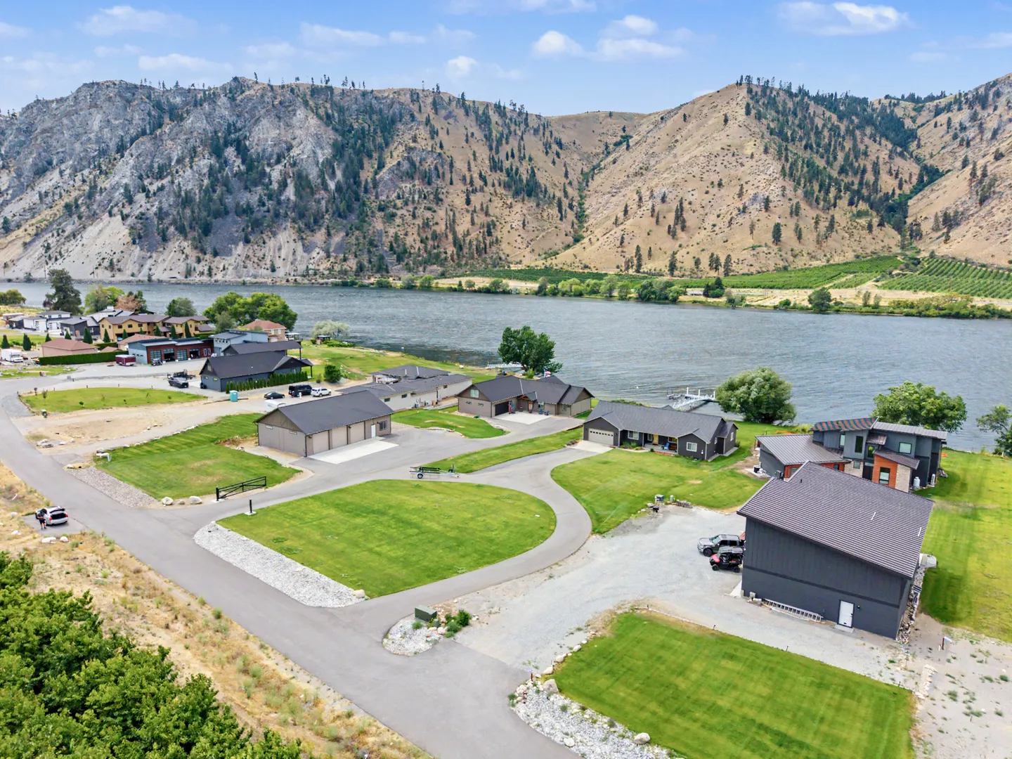 Aerial view of a waterfront neighborhood with paved driveways, modern houses, and a large river backed by rocky mountains.