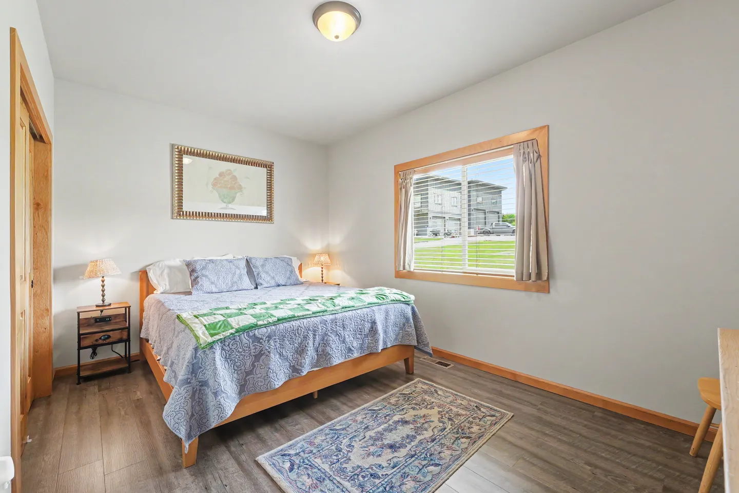 Bedroom featuring a wooden bed frame, patterned bedding, nightstand with lamp, and a window with wood trim.