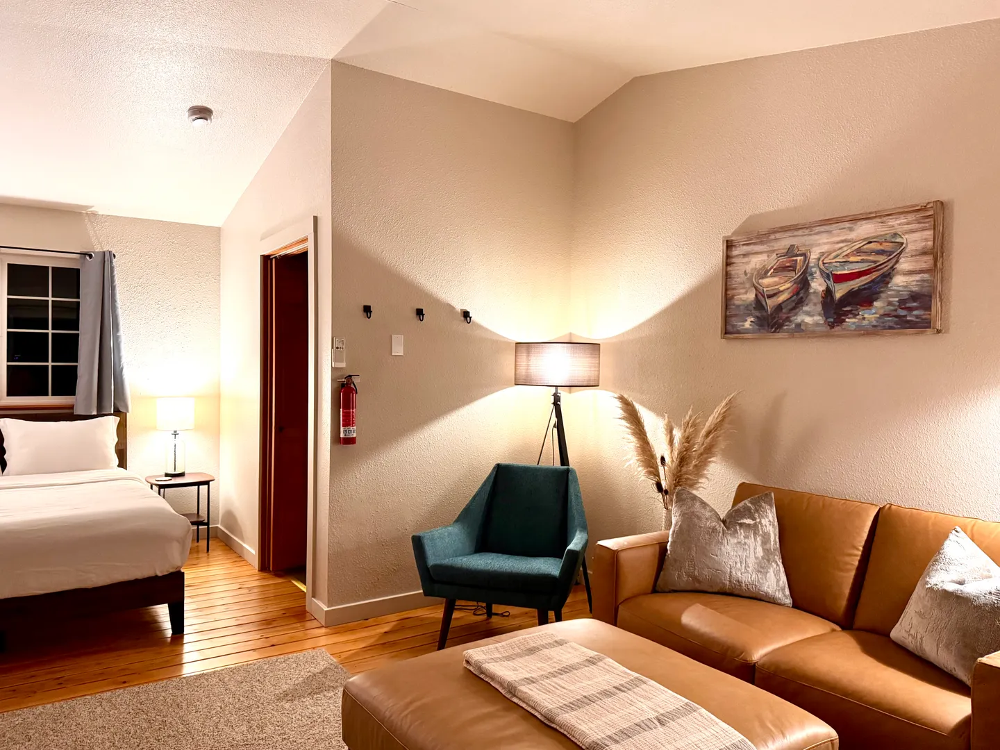 Open-concept studio interior showing a living area with a brown leather couch and teal armchair, adjacent to a bed with white linens.