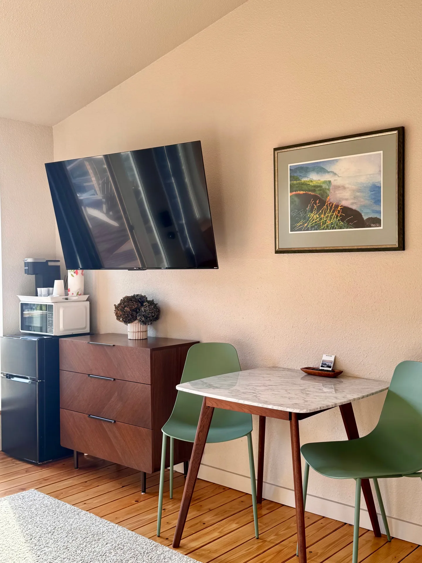 A corner seating and entertainment area with a wall-mounted TV, small dining table with green chairs, and a dresser with a mini fridge and microwave.