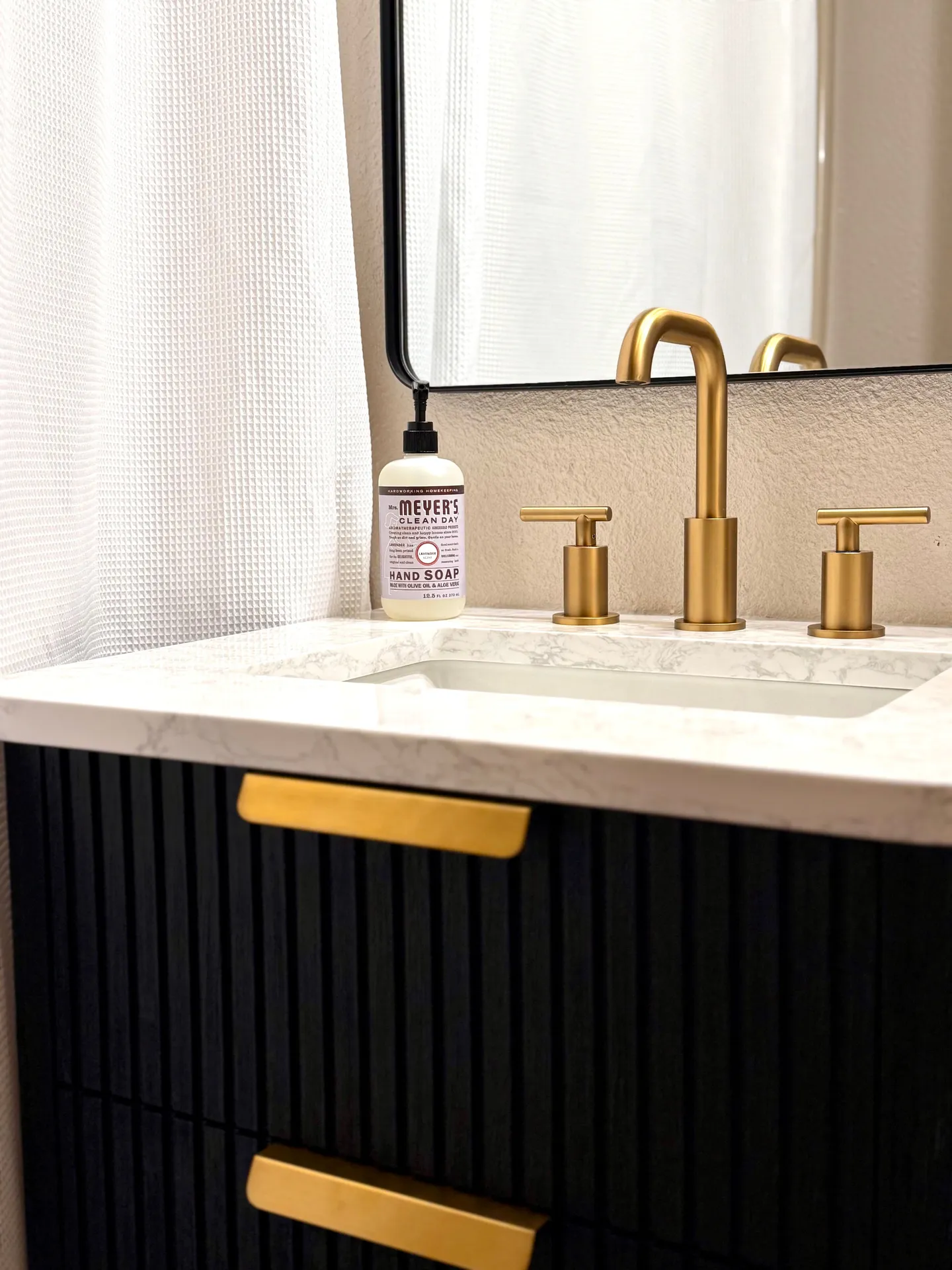 Close-up of a bathroom vanity with a black fluted cabinet, brass handles, marble-patterned countertop, brass faucet, and a soap dispenser.