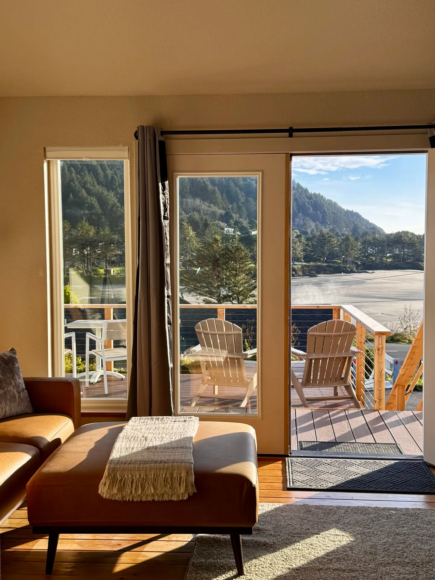 Living room looking through open doors to a sunny wooden deck with Adirondack chairs overlooking trees and a beach.