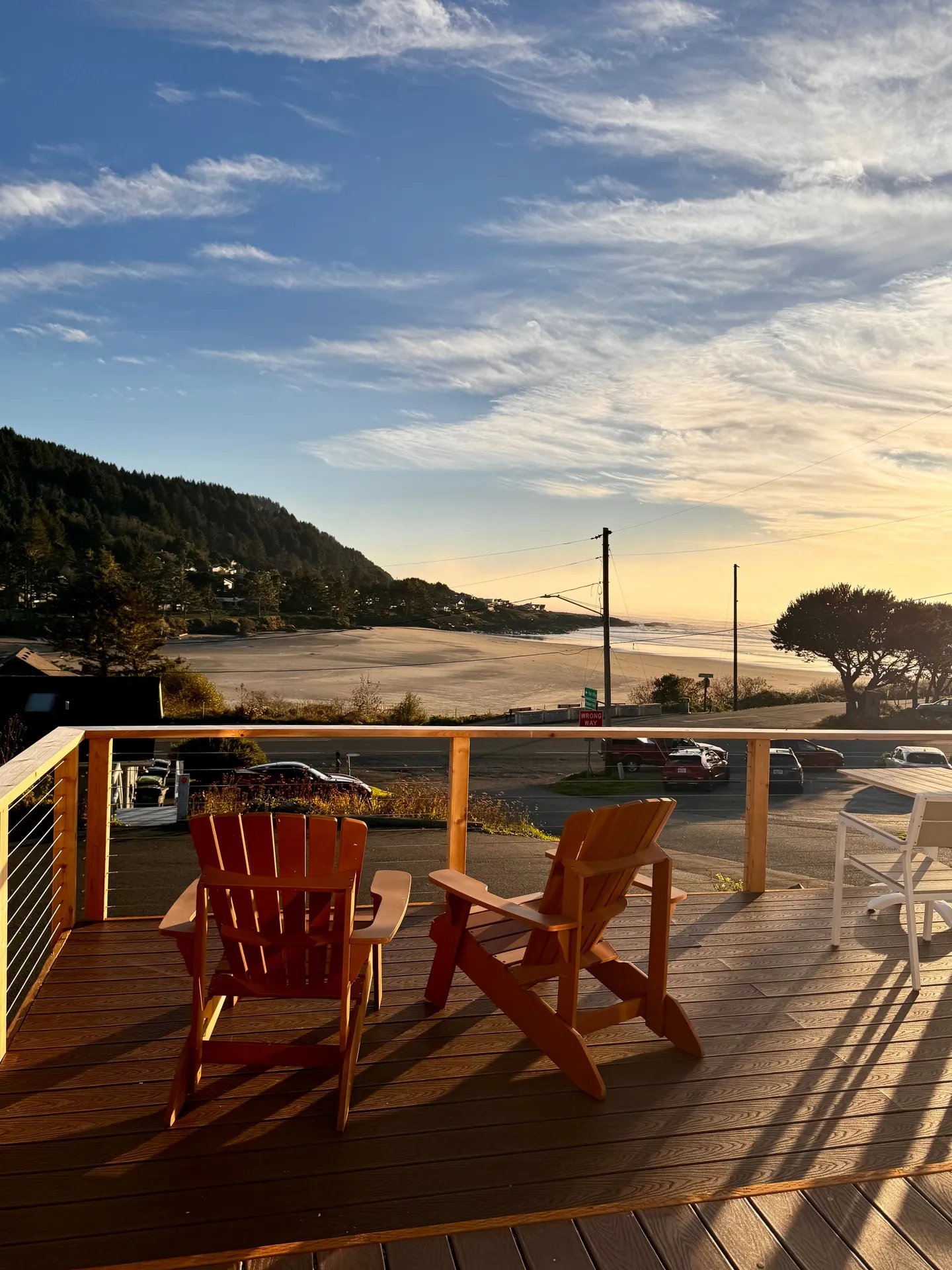 Deck with two wooden armchairs overlooking a sandy beach, forested hills, and the ocean during a sunny afternoon.