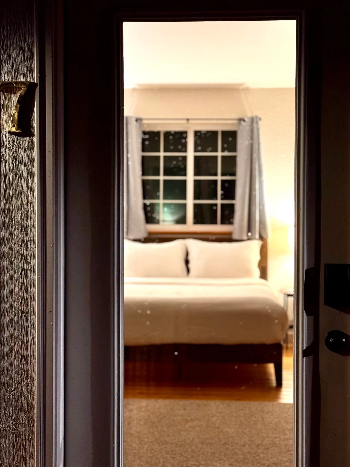 A view through an open doorway framed by a dark wall, showing a bedroom with a bed set against a paned window at night.