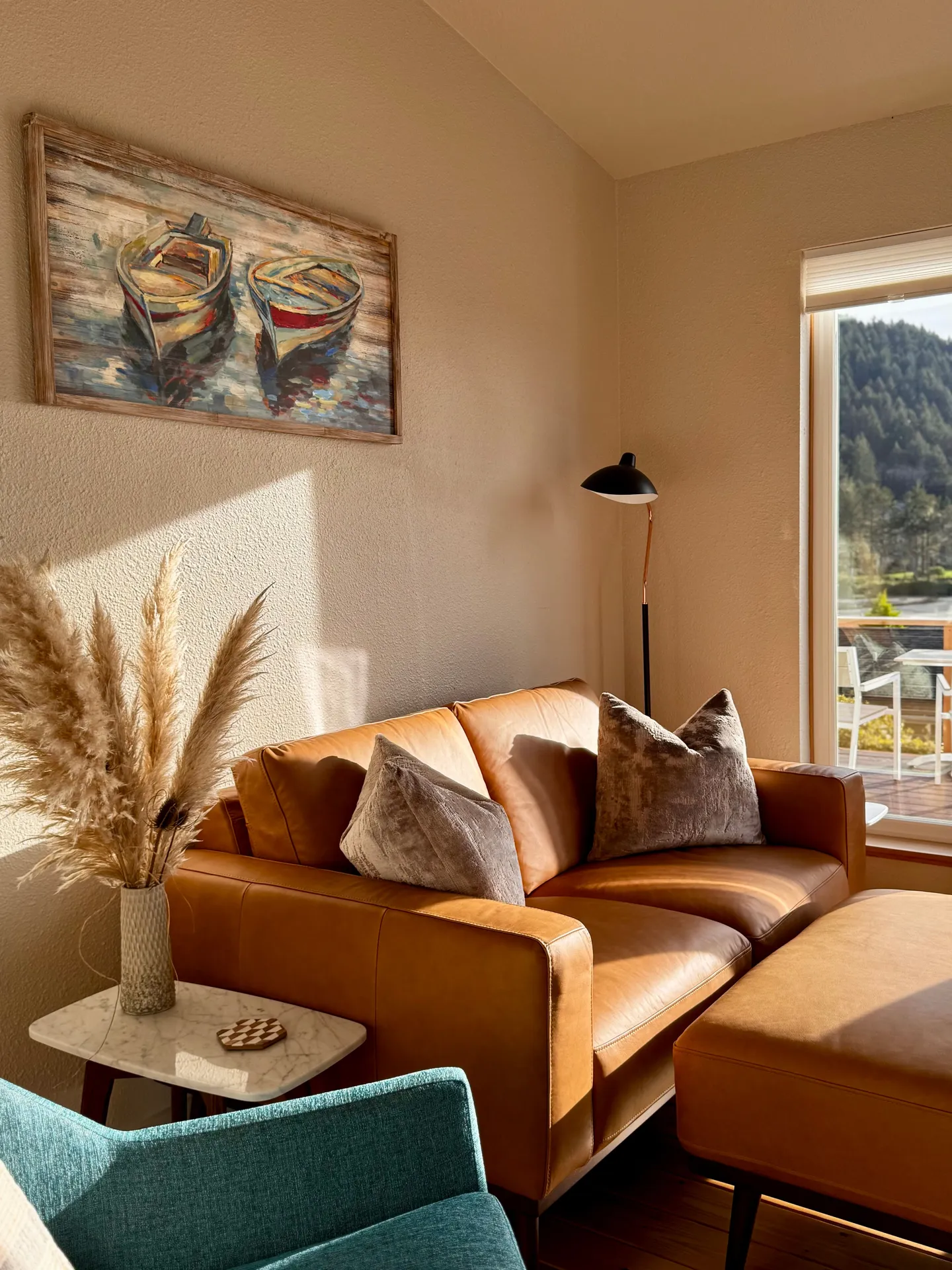 Living room corner with a tan leather loveseat, ottoman, framed boat painting, and sunlight streaming from a window.