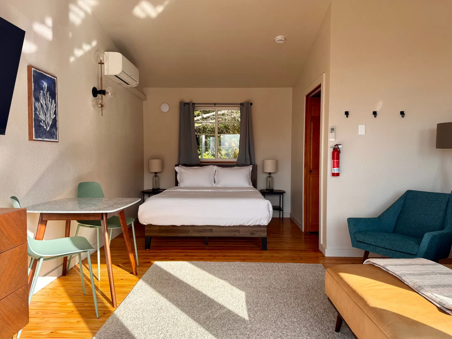 Bedroom featuring a made bed centered under a window, wood flooring, a small dining table with chairs, an armchair, and an ottoman.