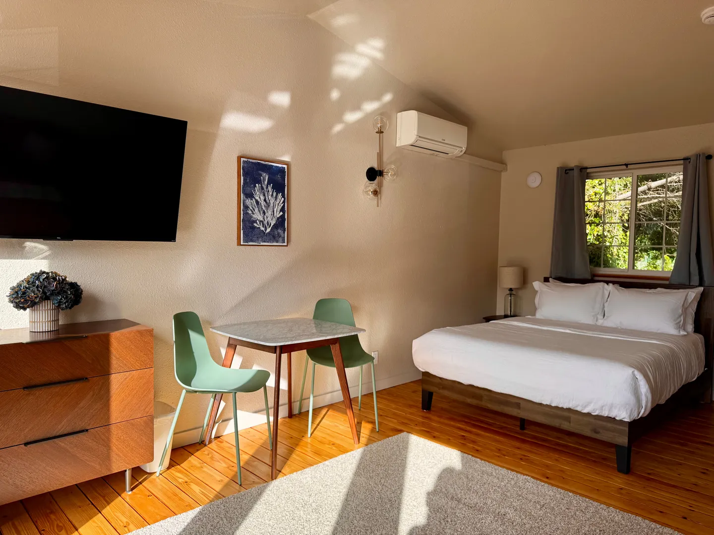 Bedroom interior featuring a bed with white linens, a small dining table with two green chairs, a wall-mounted TV, and wood floors.