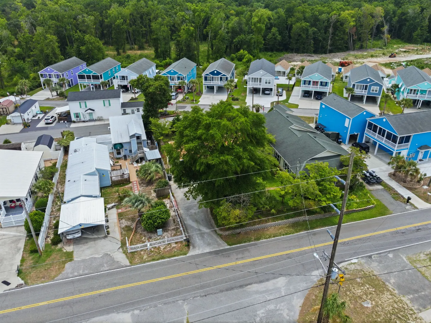 Aerial view of a coastal neighborhood with single-family houses, paved road, and dense green trees in the background.