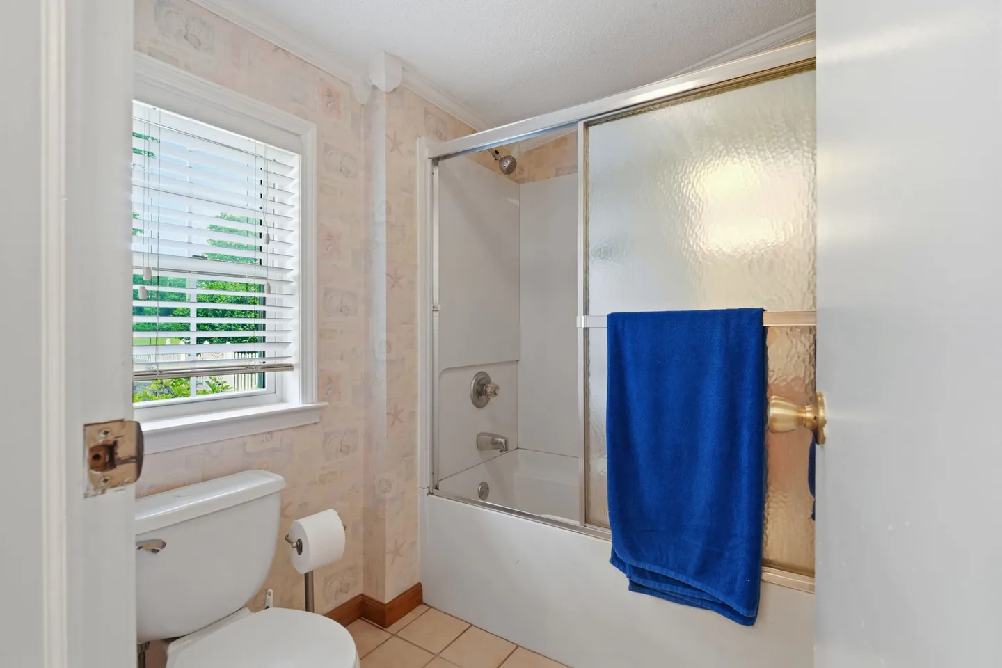 Bathroom showing a white toilet, tiled floor, a window with horizontal blinds, and a bathtub with sliding frosted glass doors.