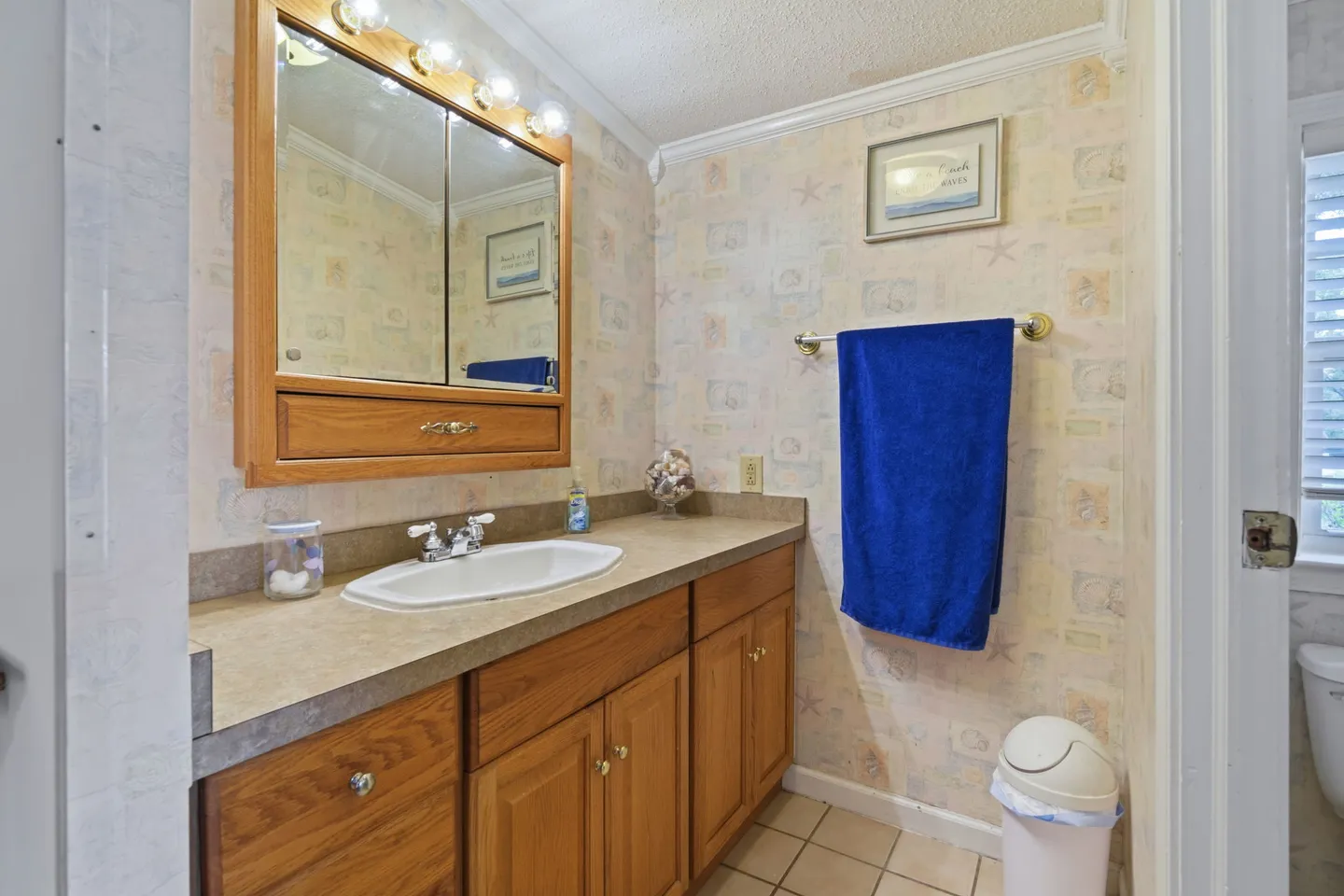 Bathroom with oak vanity, sink, framed medicine cabinet mirror with light bulbs, and sea-themed wallpaper.