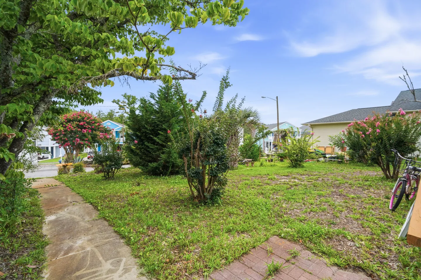 Outdoor view of a green grassy yard with trees, flowering bushes, a paved walkway, and neighboring houses under a blue sky.