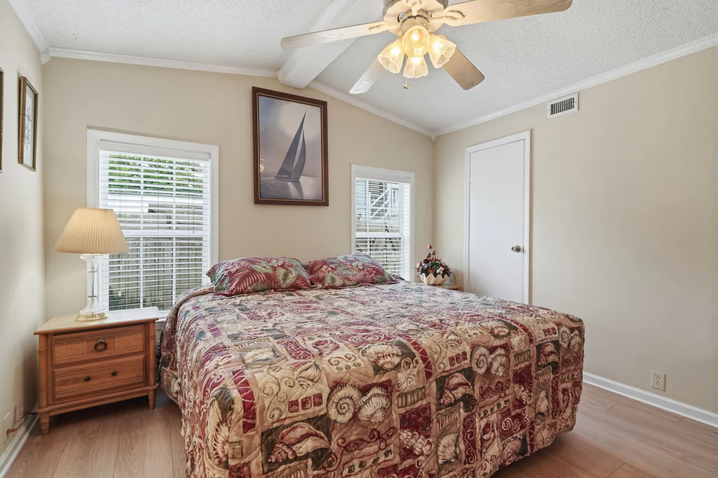 Bedroom with a patterned bedspread, wooden nightstand, two windows with blinds, sailboat artwork, and a ceiling fan.