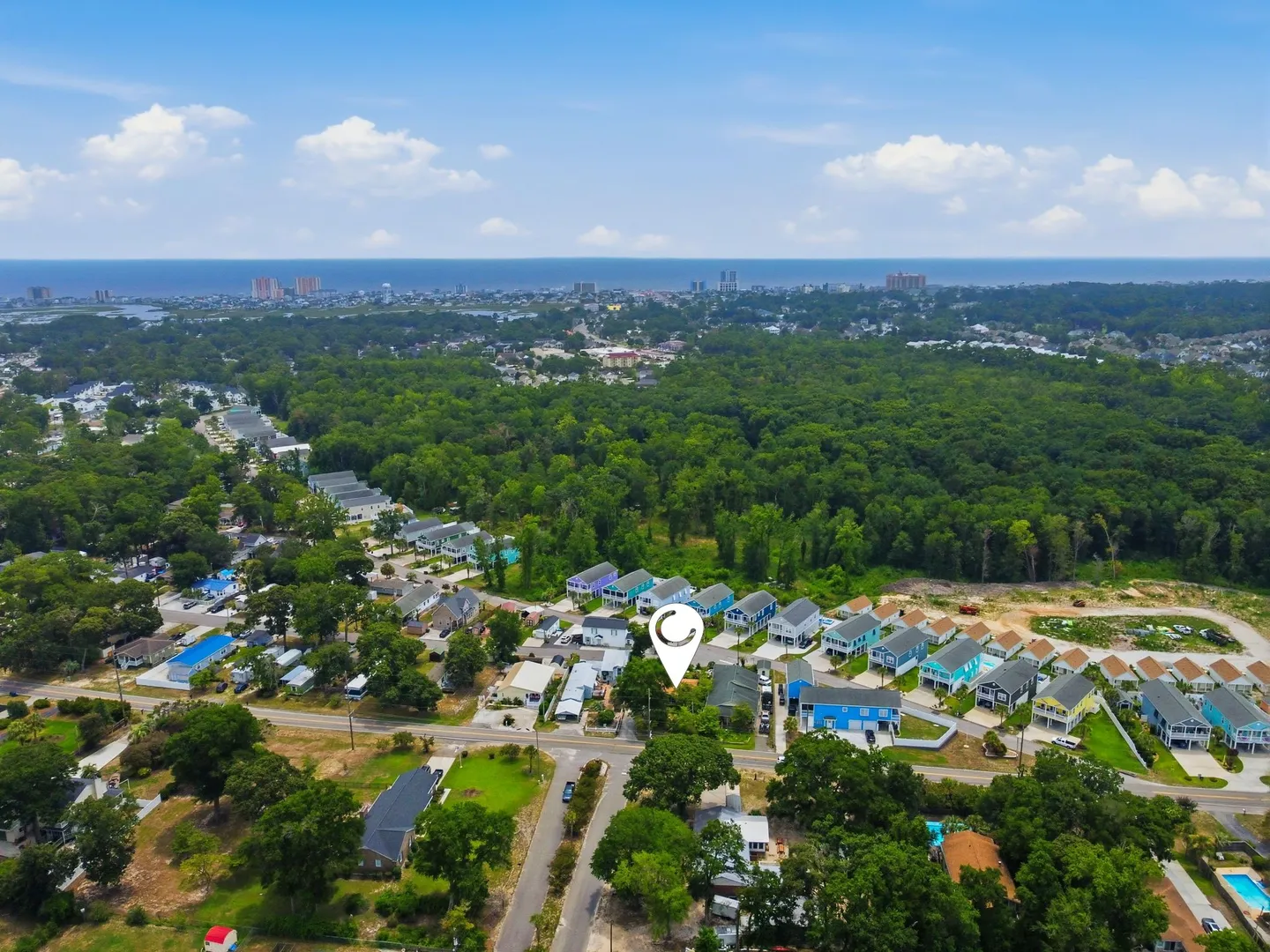 High aerial view of a coastal neighborhood with dense green trees, colorful houses, and the ocean visible on the horizon under a partly cloudy sky.
