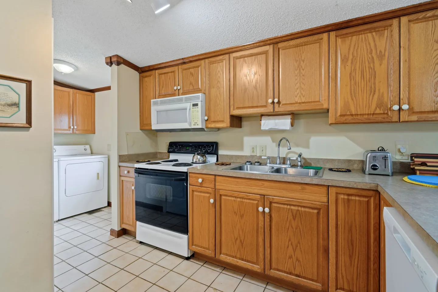 Kitchen with wooden cabinetry, white appliances, double sink, and tiled floor, with laundry appliances visible in an adjacent space.
