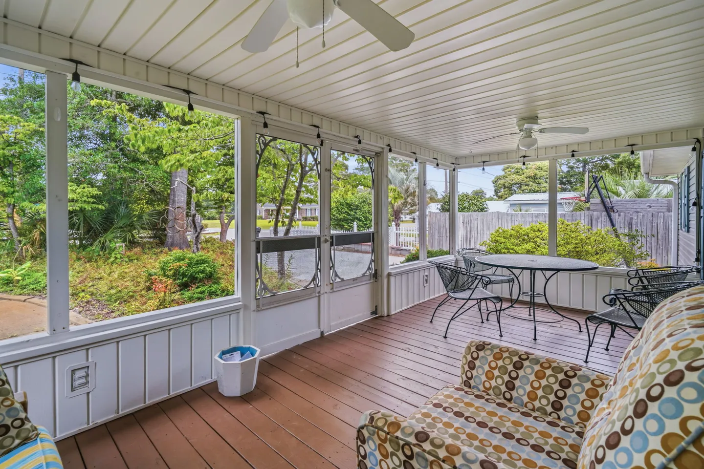 Screened-in porch with wood-plank flooring, ceiling fans, patterned armchairs, and a metal dining table set overlooking a fenced yard.