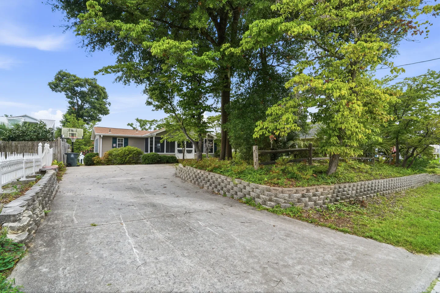 A wide concrete driveway leads toward a single-story home with a screened front porch, flanked by stone retaining walls and mature trees under a blue sky.