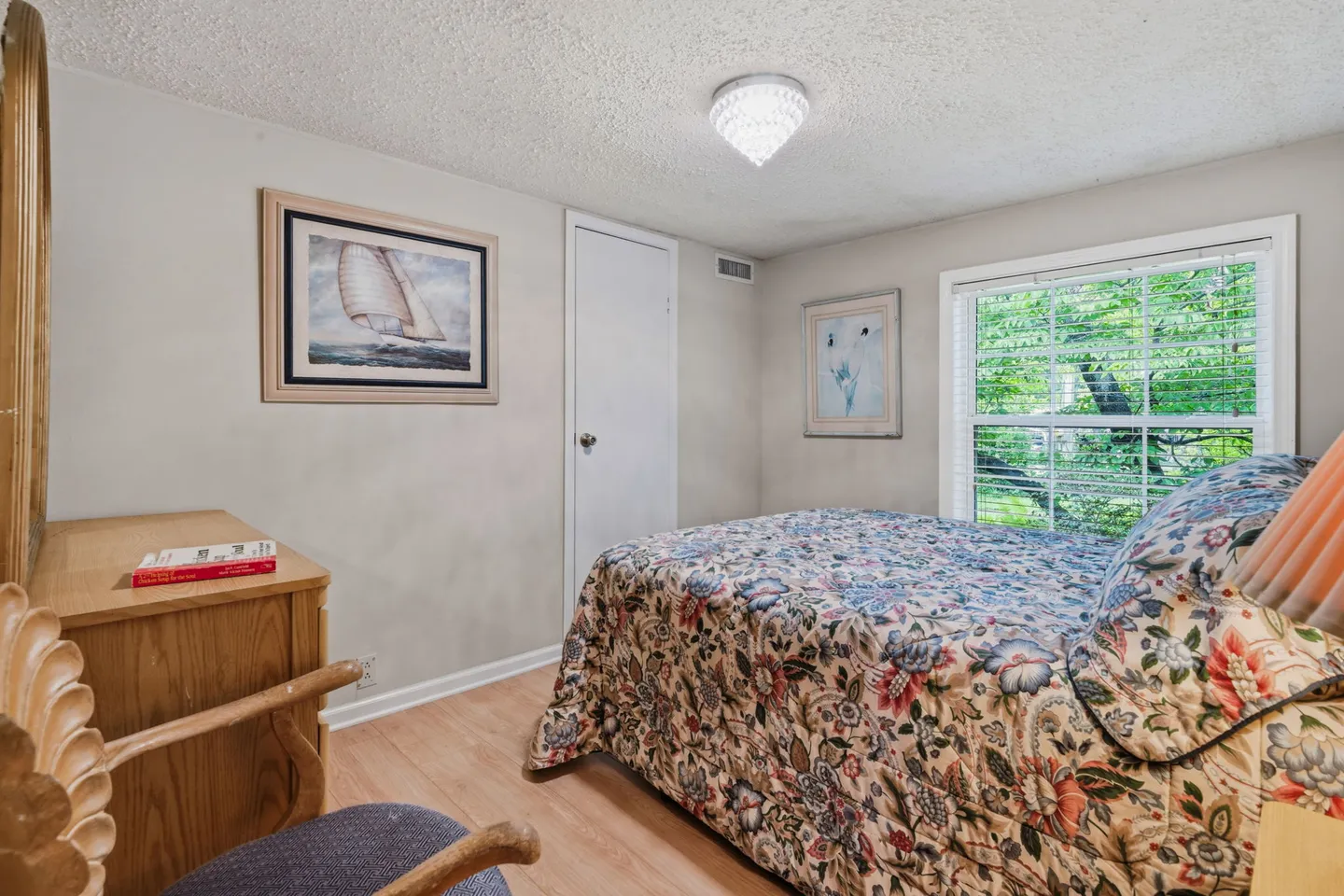 Bedroom featuring a floral patterned bedspread, wood-look flooring, light grey walls, and a window with blinds looking out to green foliage.