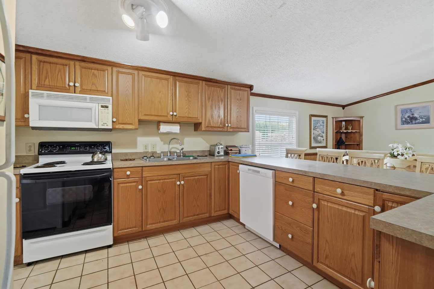 A kitchen with wood cabinets, tiled floor, a white electric range, and a breakfast bar overlooking a dining area.