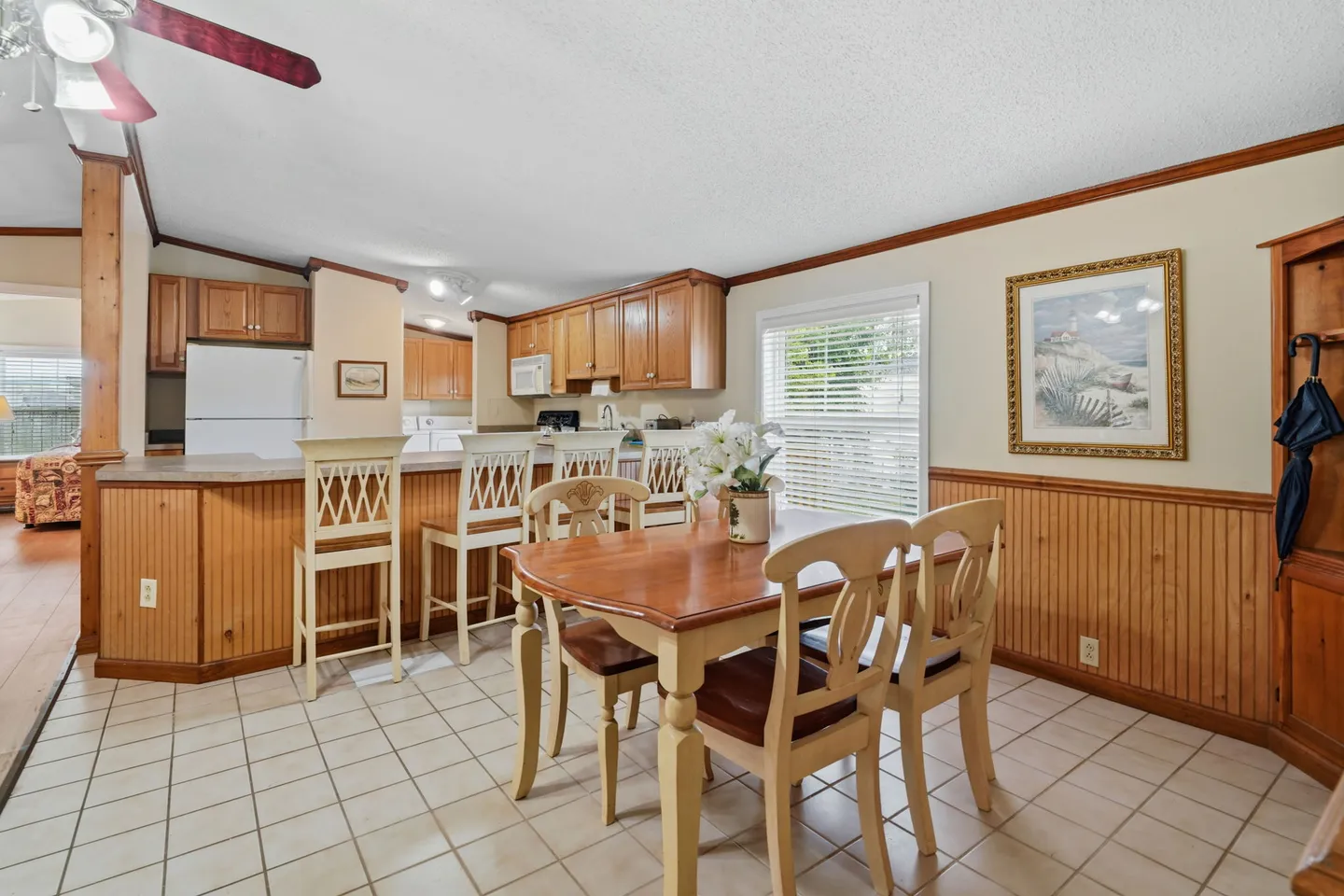 Dining area with a wooden table and chairs adjacent to a kitchen island with bar stools, featuring tile flooring and wood wainscoting.