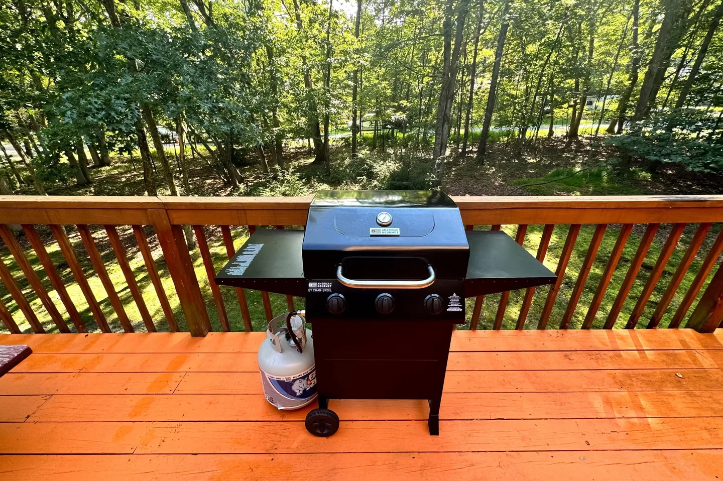 A black gas grill with a propane tank on an orange wooden deck overlooking dense green trees.