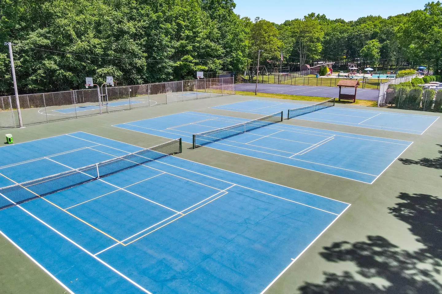 Aerial view of community outdoor sports facilities with multiple blue tennis courts, pickleball lines, and a fenced basketball court surrounded by trees.