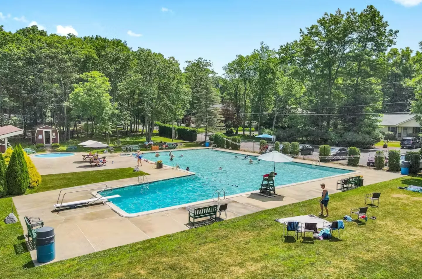 An elevated view of an outdoor community swimming pool surrounded by a concrete deck, lawn, and dense green trees.