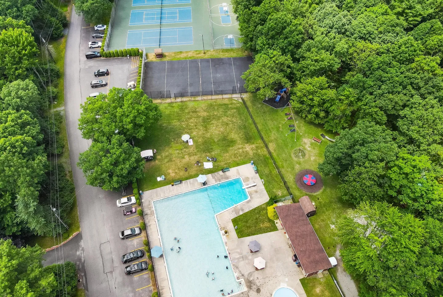 Aerial view of a community recreation area with a large swimming pool, tennis and basketball courts, parking areas, and surrounding woods.