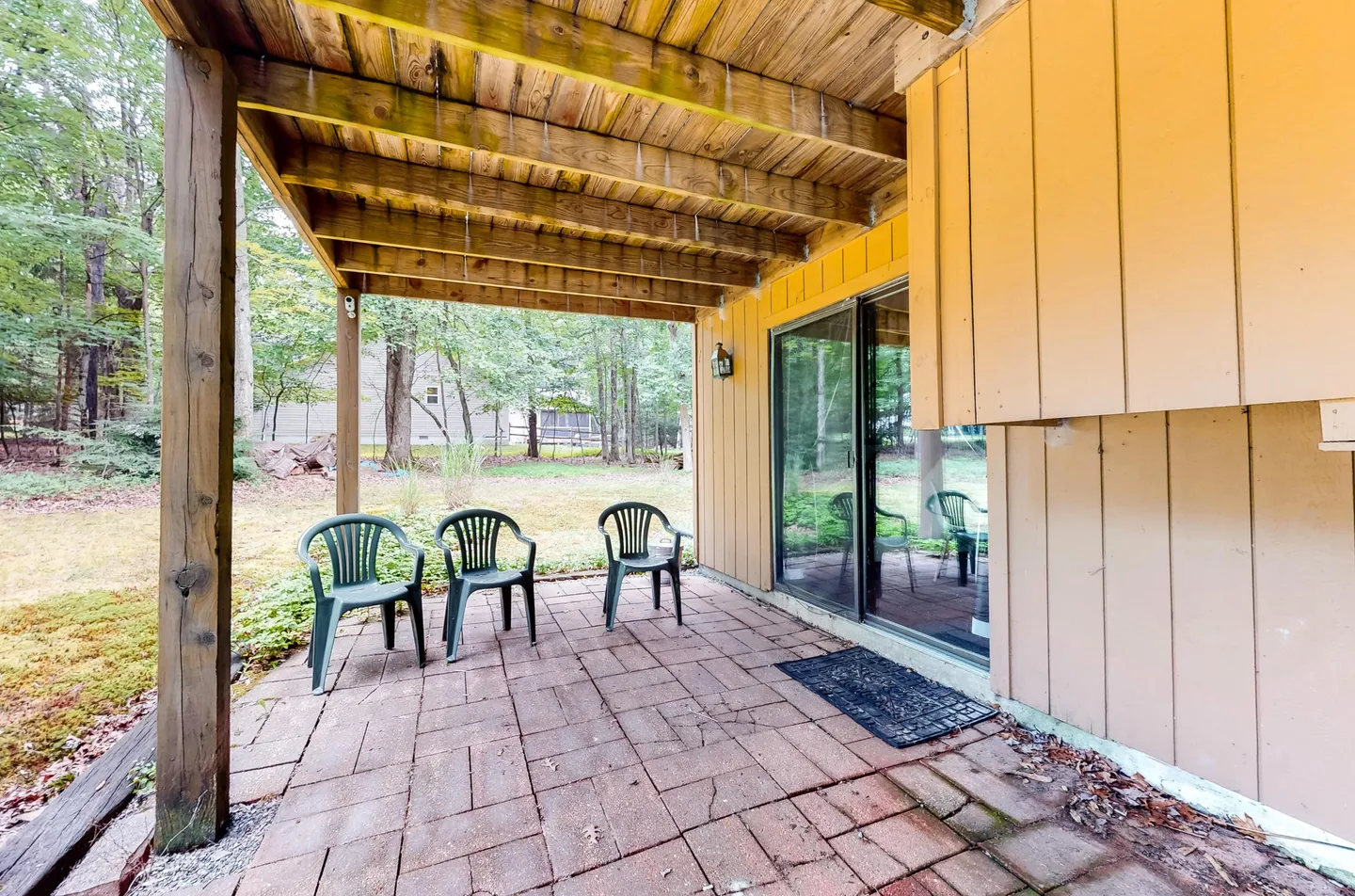 A covered brick paver patio beneath an upper deck, featuring three green plastic chairs and sliding glass doors leading indoors.