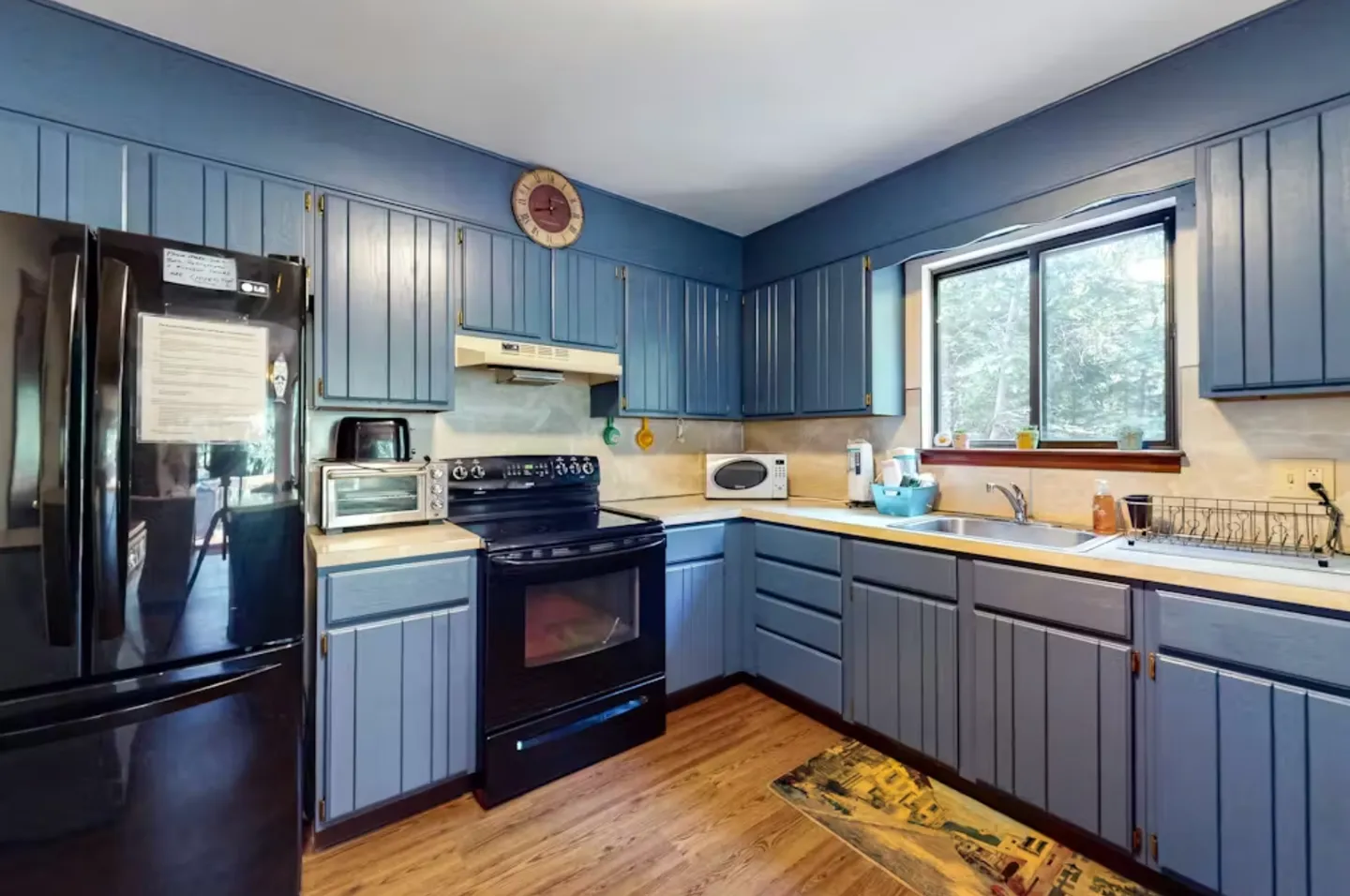 Kitchen with blue-painted paneled cabinetry, light countertops, black appliances, and a window above the sink.
