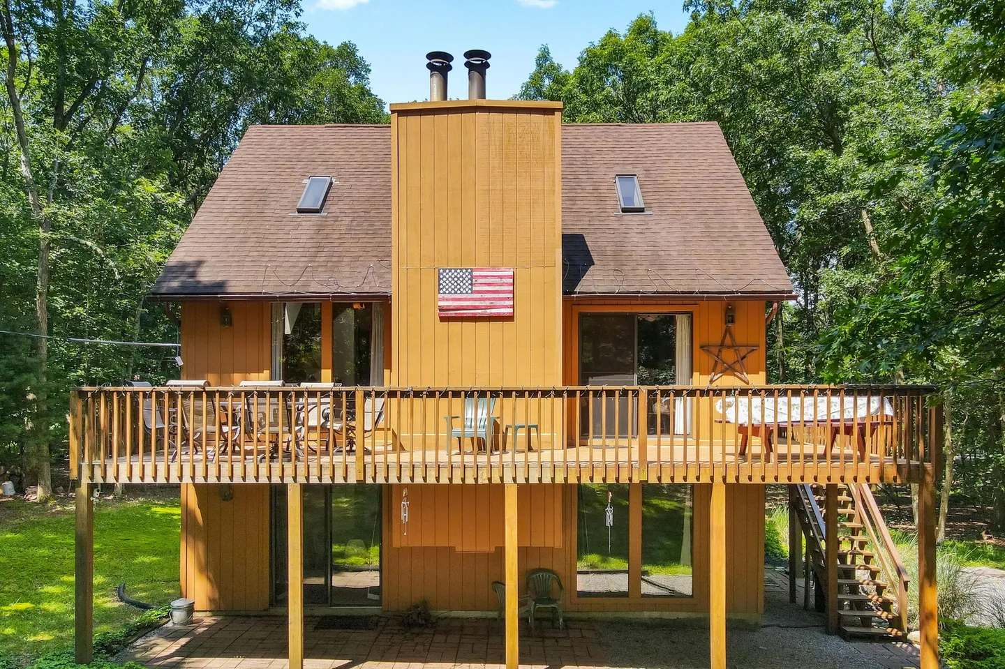Rear exterior view of a two-story wood-sided house with an elevated deck, patio below, and surrounding woods.