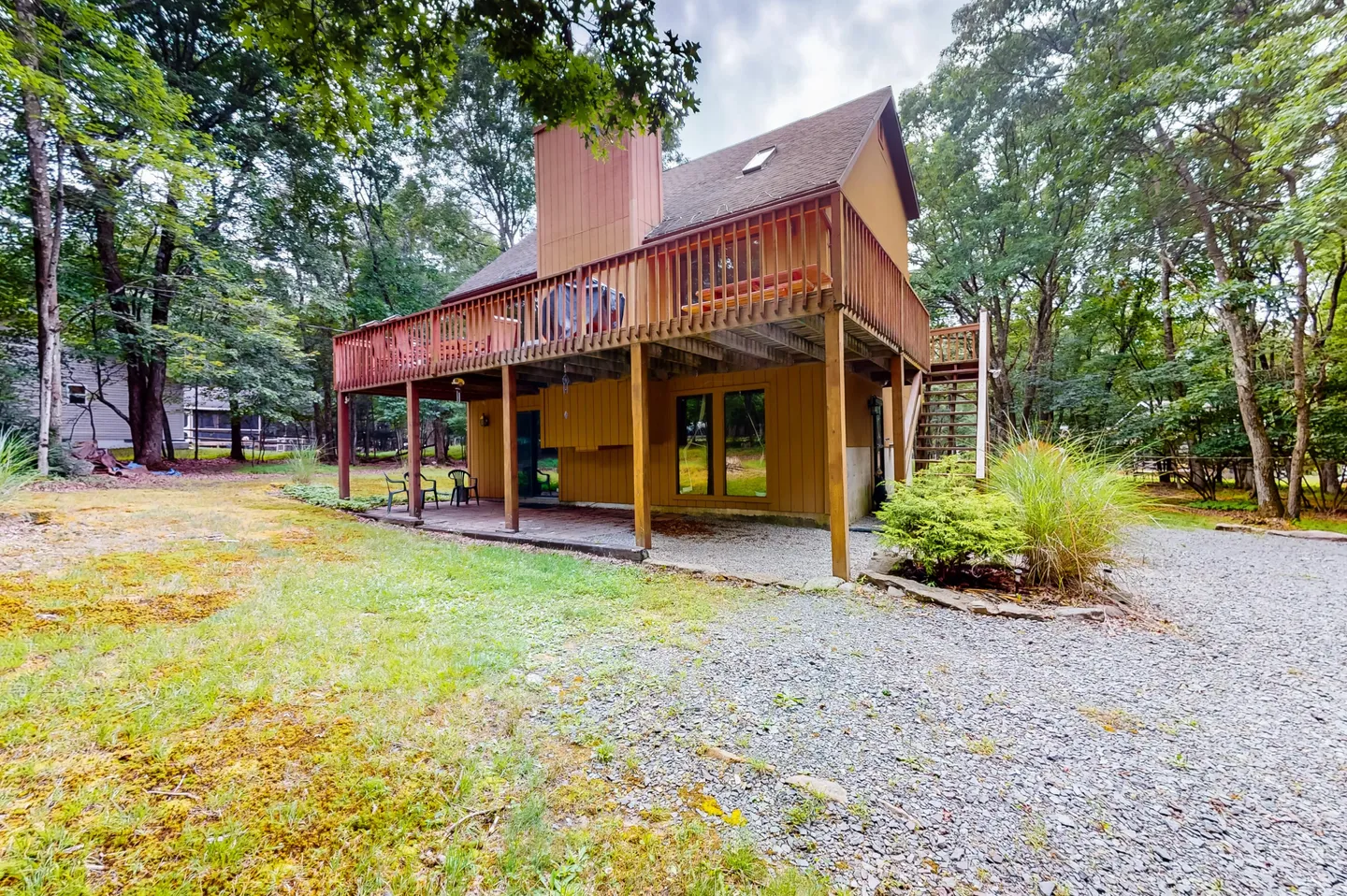 Rear exterior view of a multi-story wood-sided house with a large elevated wooden deck, lower patio area, and gravel yard surrounded by trees.