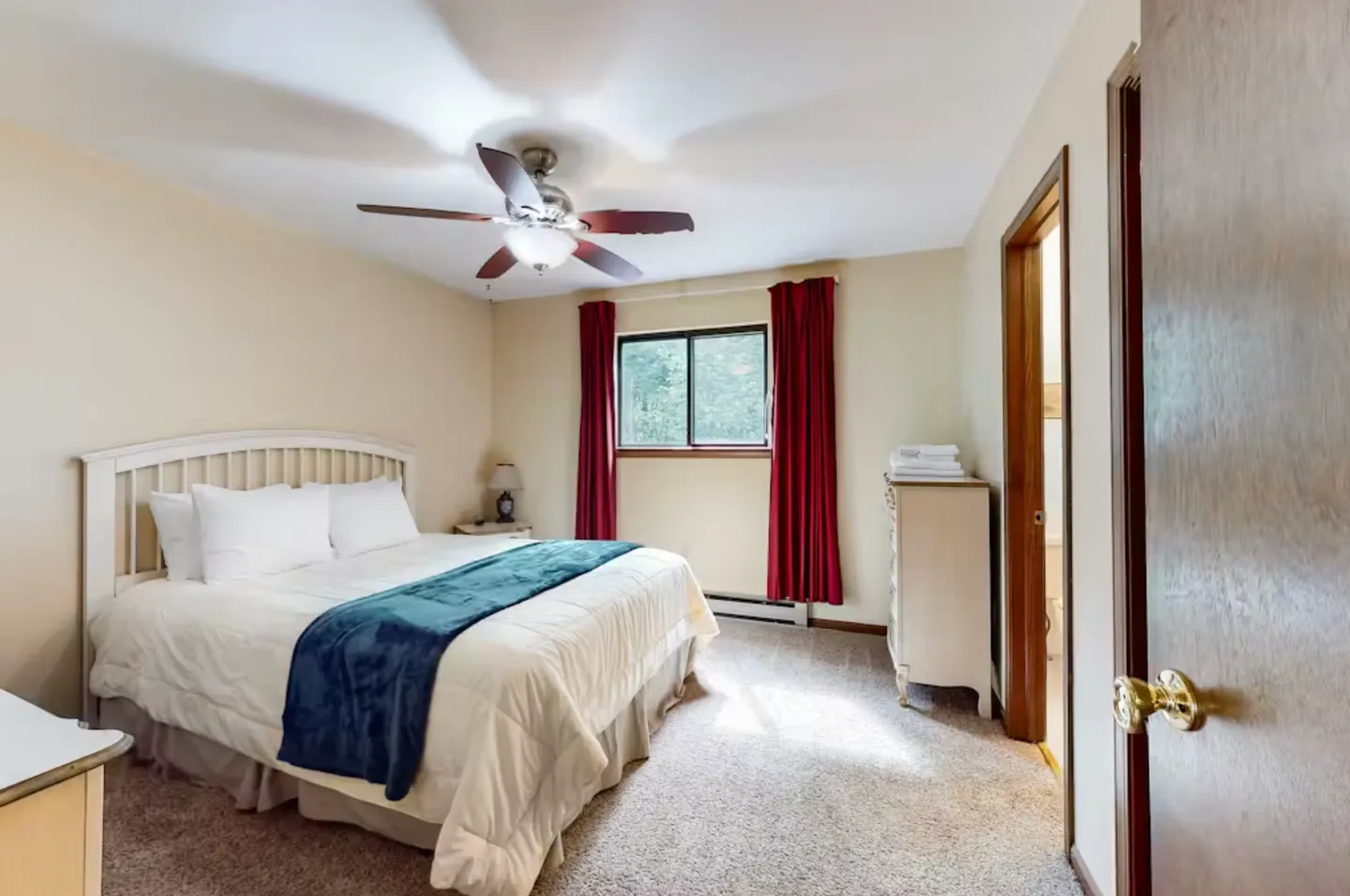 Bedroom with a white headboard, made bed, carpeted flooring, ceiling fan, and red curtains framing a window.