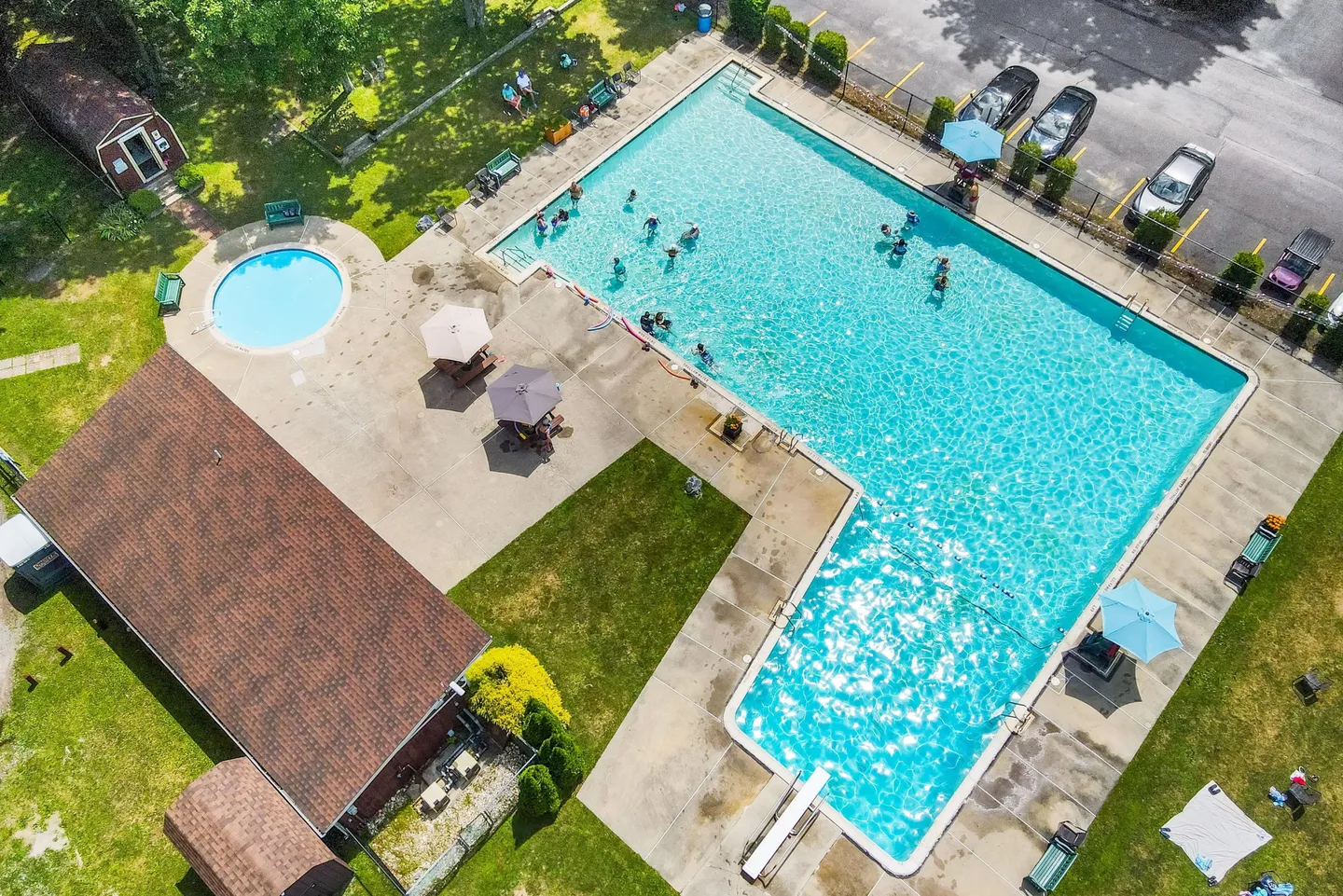 Aerial view of an L-shaped outdoor swimming pool with swimmers, a separate round wading pool, sun umbrellas, and adjacent buildings.