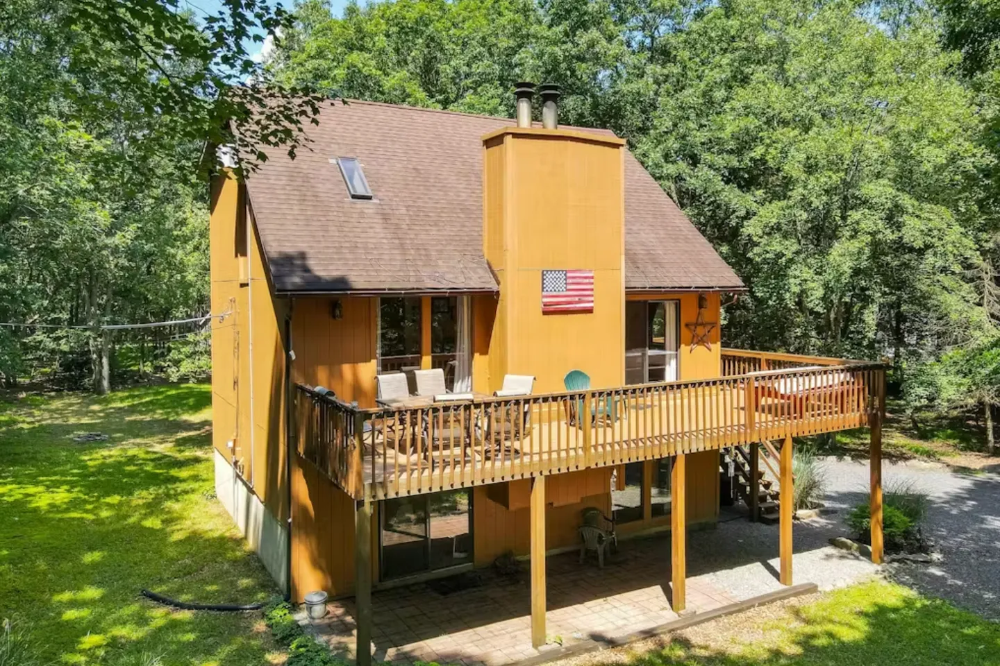 Exterior elevated view of a brown two-story house with an elevated wooden deck, patio underneath, and surrounding trees.