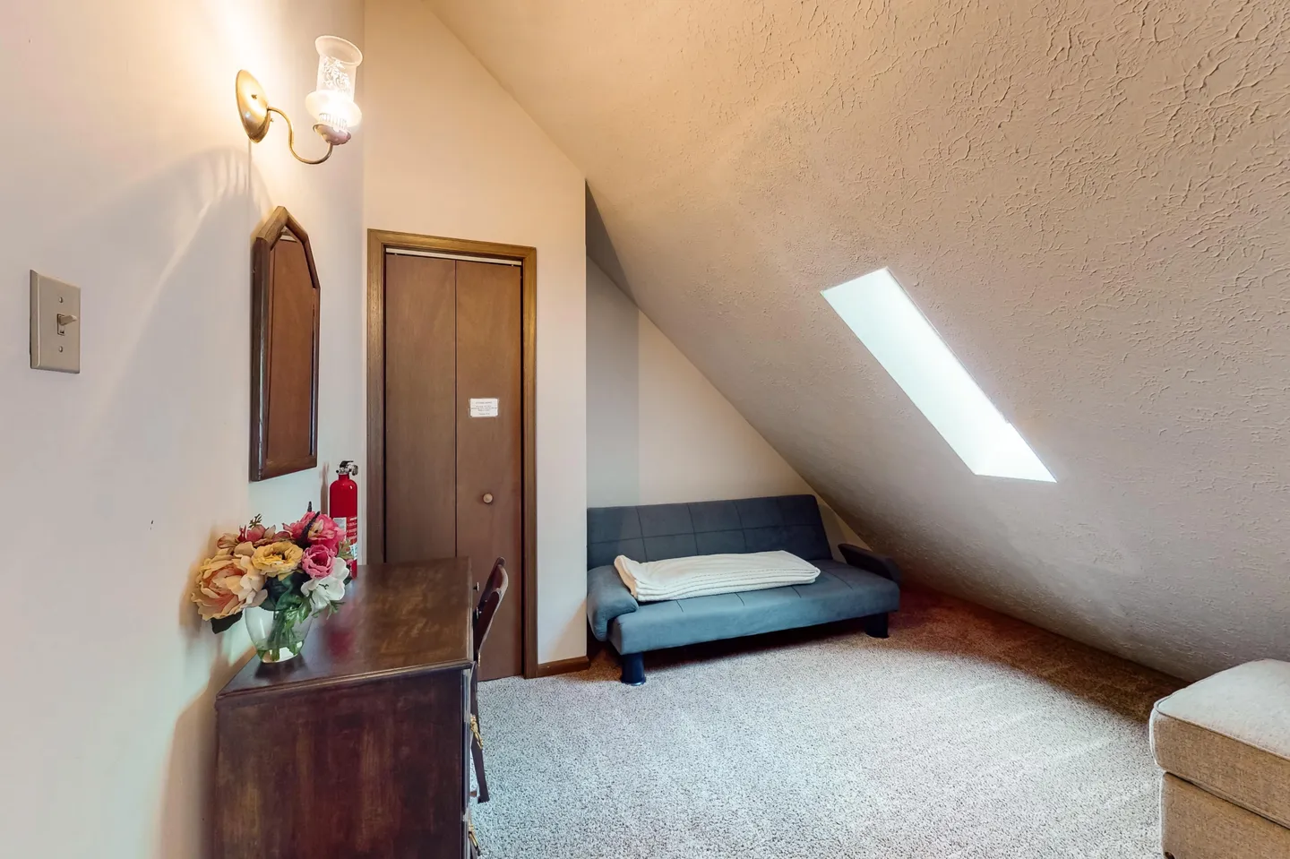 Attic-style room with sloped ceiling, skylight, carpet, a futon sofa, a vanity desk, and a closet door.