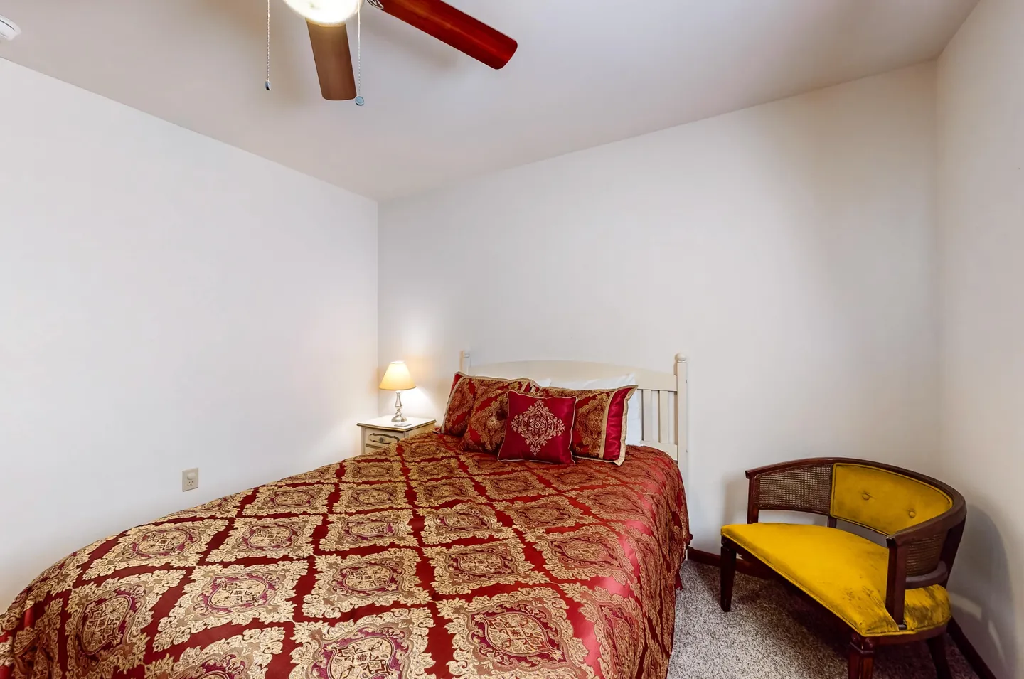 Bedroom featuring a bed with ornate red and gold bedding, a white headboard, a small bedside table with lamp, a ceiling fan, and a yellow cushioned chair.