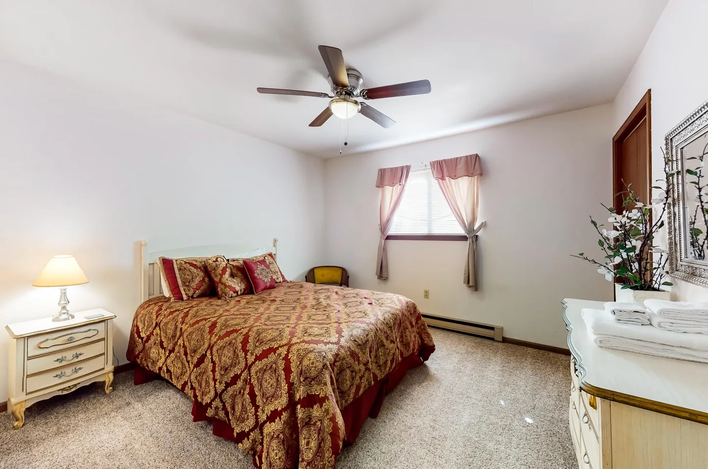 Bedroom with patterned red and gold bedspread, bedside table with lamp, ceiling fan, and dresser.