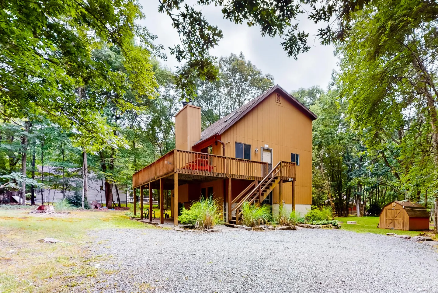 Exterior view of a two-story wood-sided cabin with an elevated wrap-around deck, outdoor stairs, gravel driveway, and surrounding trees.