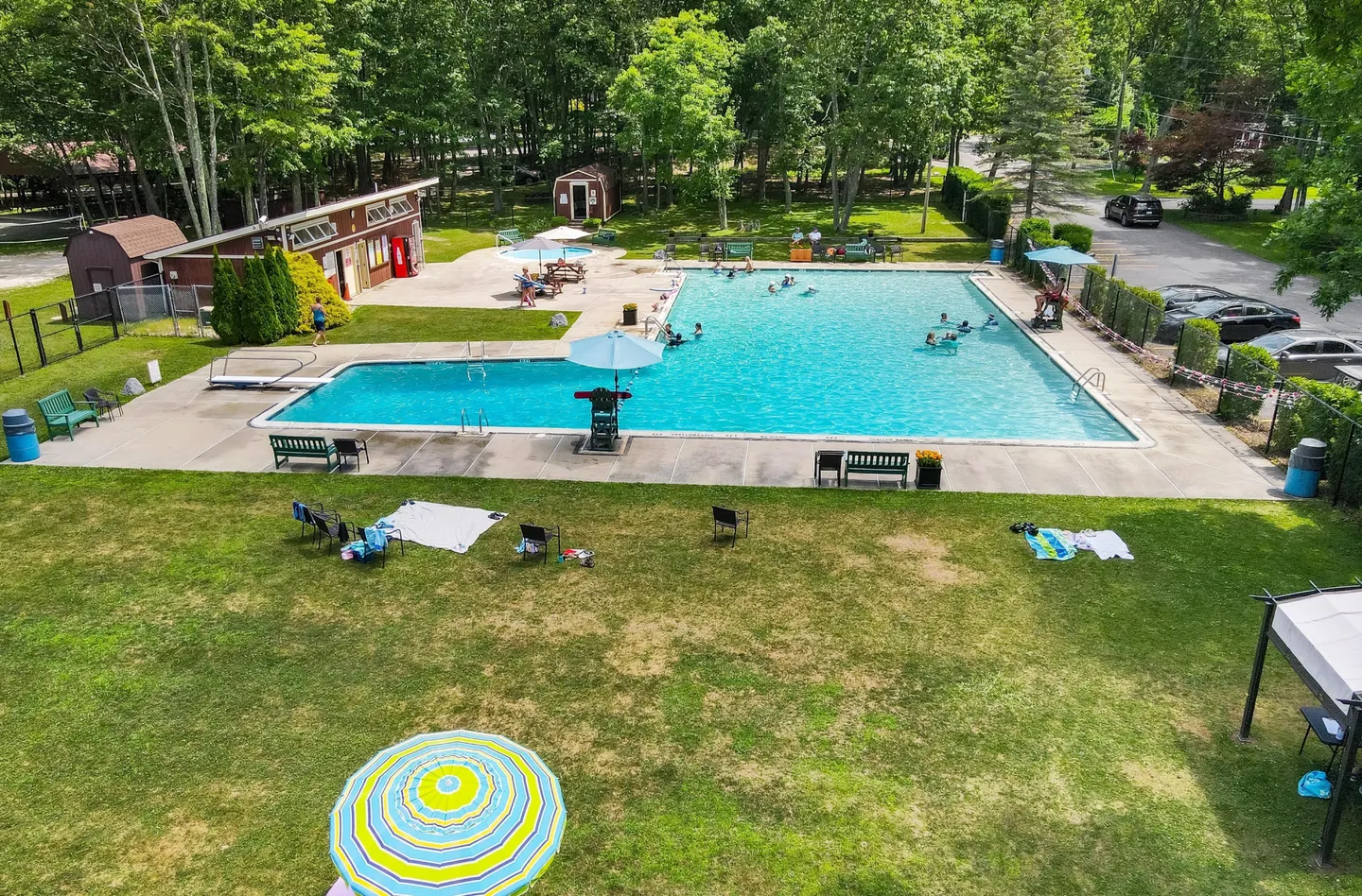 Elevated view of a large outdoor swimming pool surrounded by a concrete deck, green lawn, and trees.