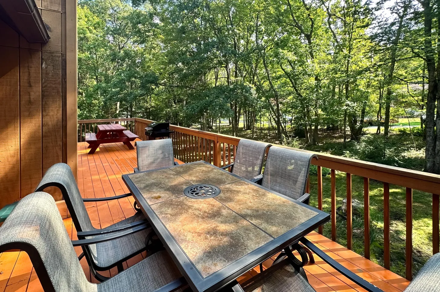 Outdoor wooden deck featuring a dining table with chairs, an adjacent picnic table, and a barbecue grill surrounded by dense green woods.