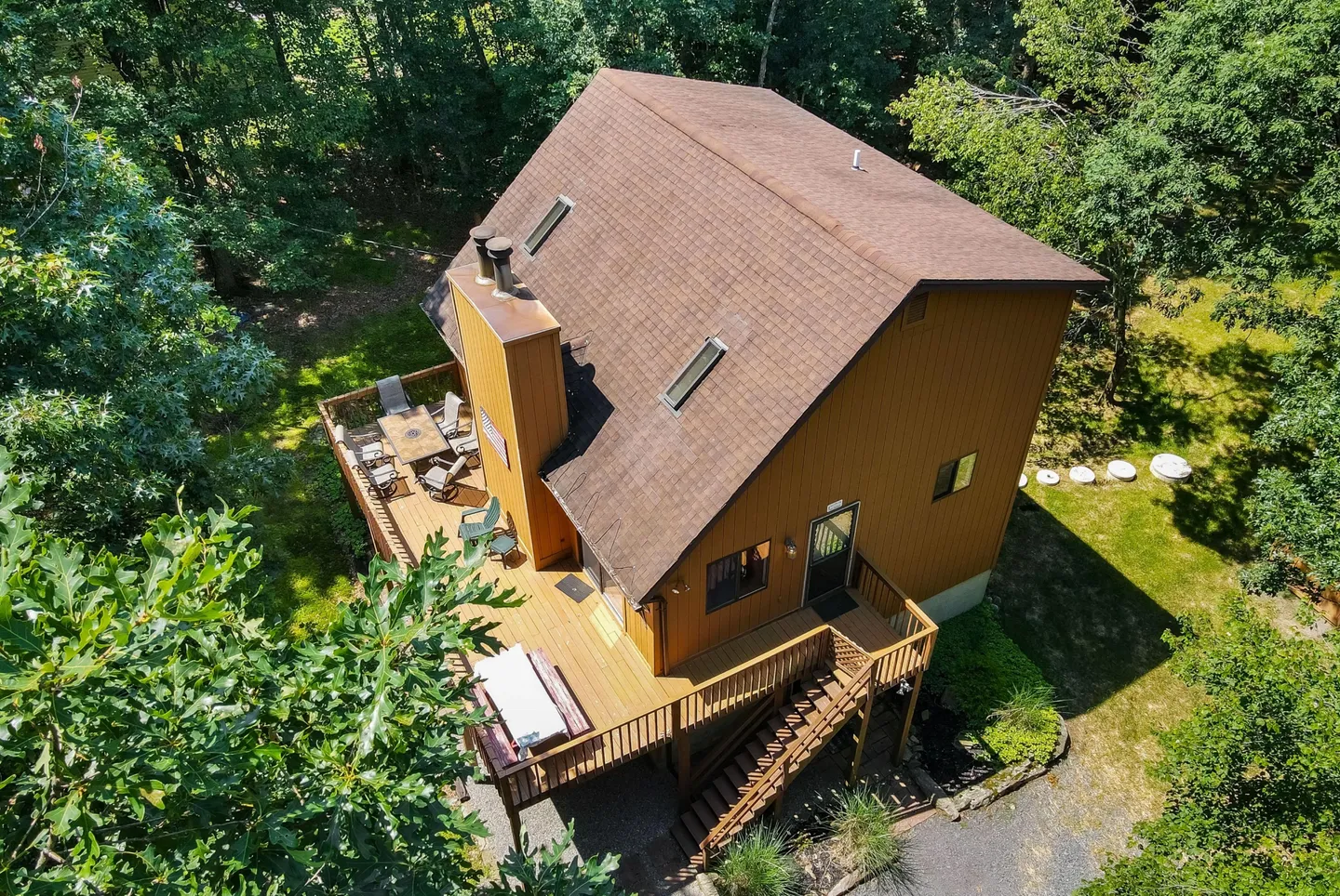 High-angle view of a brown wooden cabin surrounded by lush green trees, featuring a multi-level back deck with patio furniture and a hot tub.