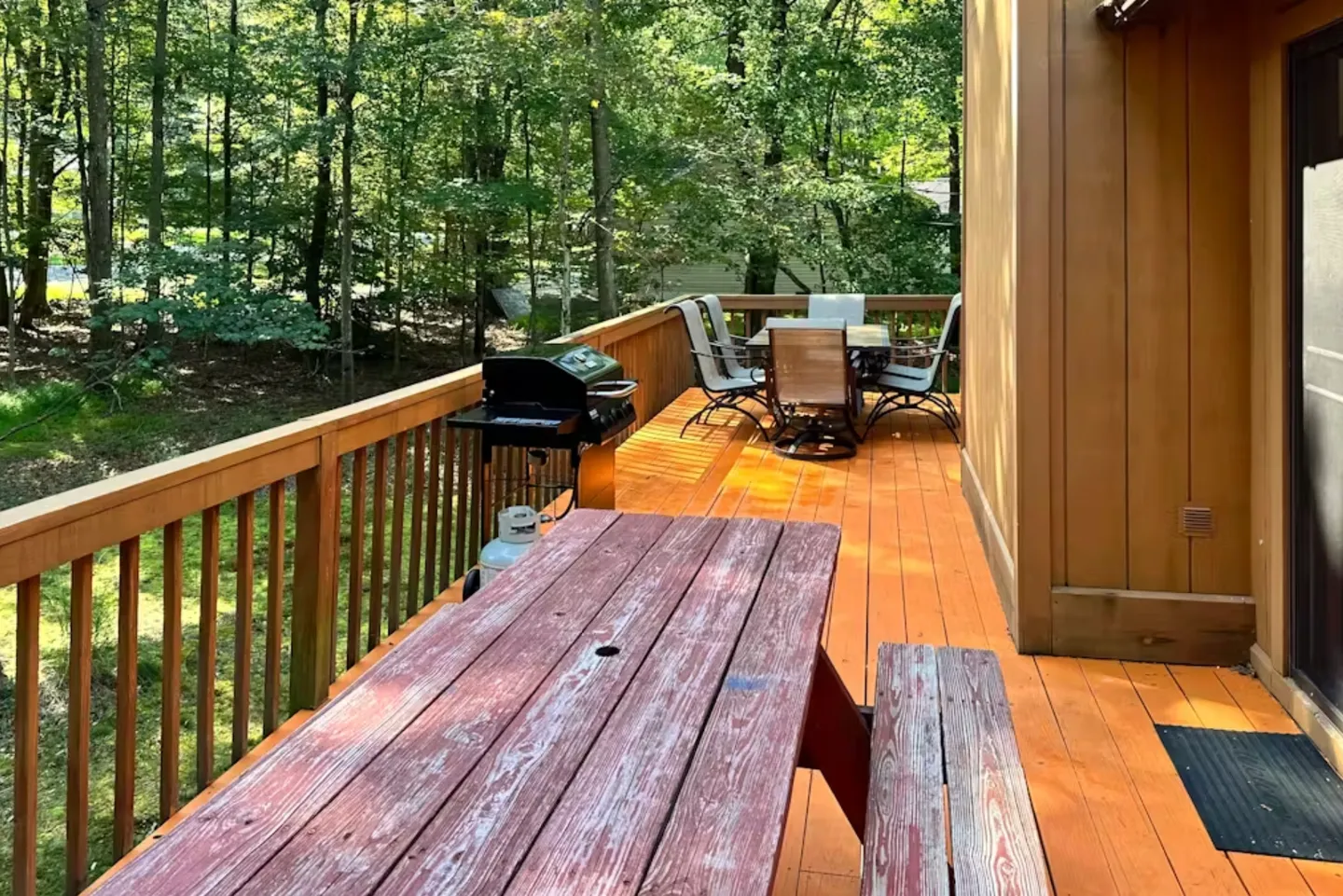 Outdoor wooden deck with a picnic table, gas grill, and patio dining set surrounded by trees.