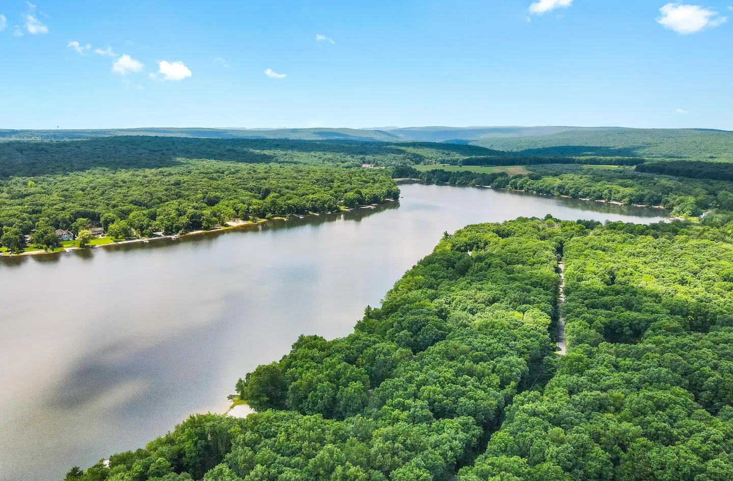 Aerial view of a wide lake surrounded by dense green forest under a partly cloudy blue sky.