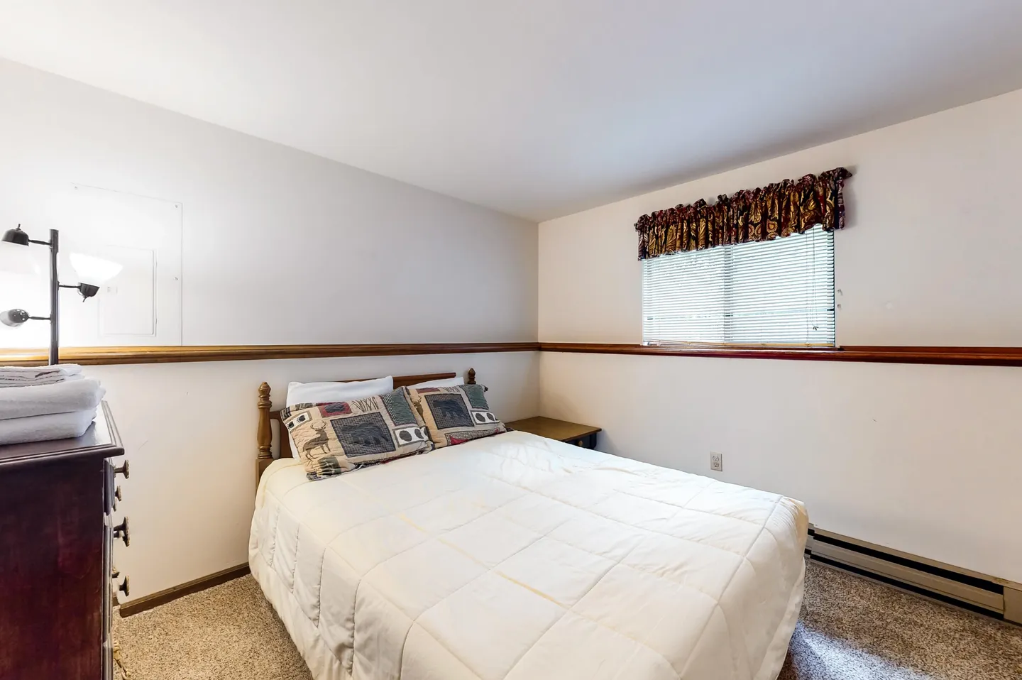 Bedroom with a white comforter on a bed, carpet flooring, a window with horizontal blinds, and a wooden ledge running along the wall.