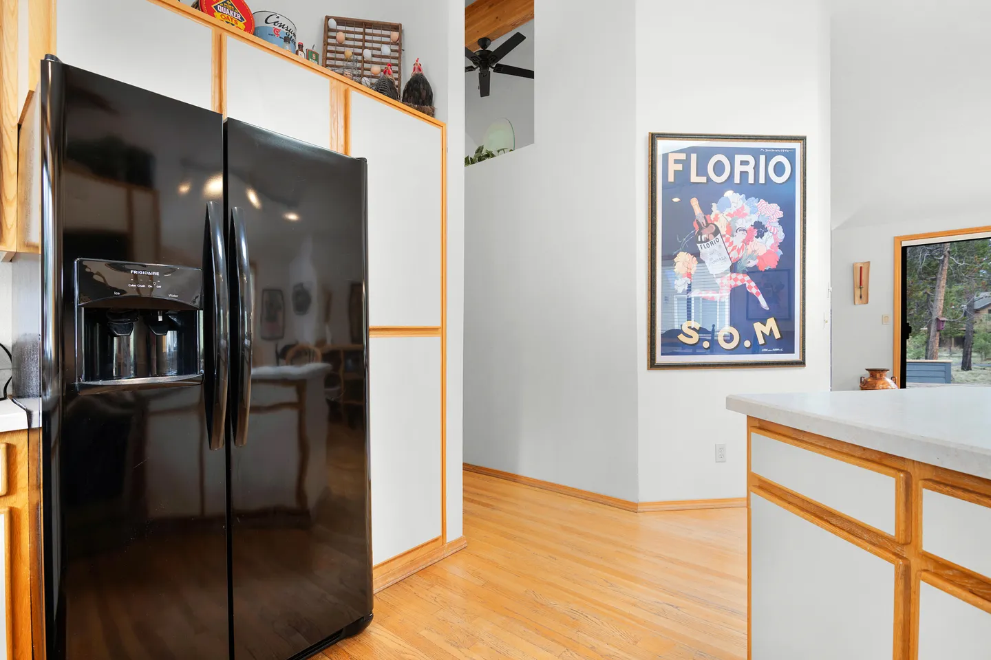 Kitchen view showing a black double-door refrigerator, wood-trimmed white cabinets, light wood flooring, and a framed Florio poster on the hallway wall.