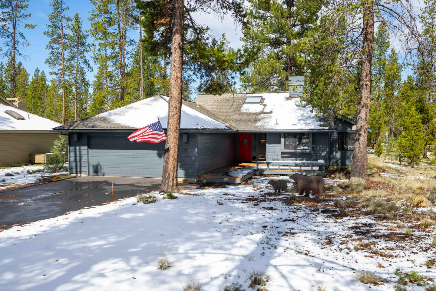 Exterior front view of a grey single-story house surrounded by tall pine trees and light snow on the ground and roof.