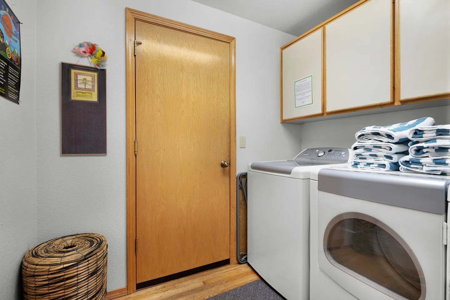 Laundry room with a washing machine, dryer, upper cabinets, a woven hamper, and an interior wooden door.