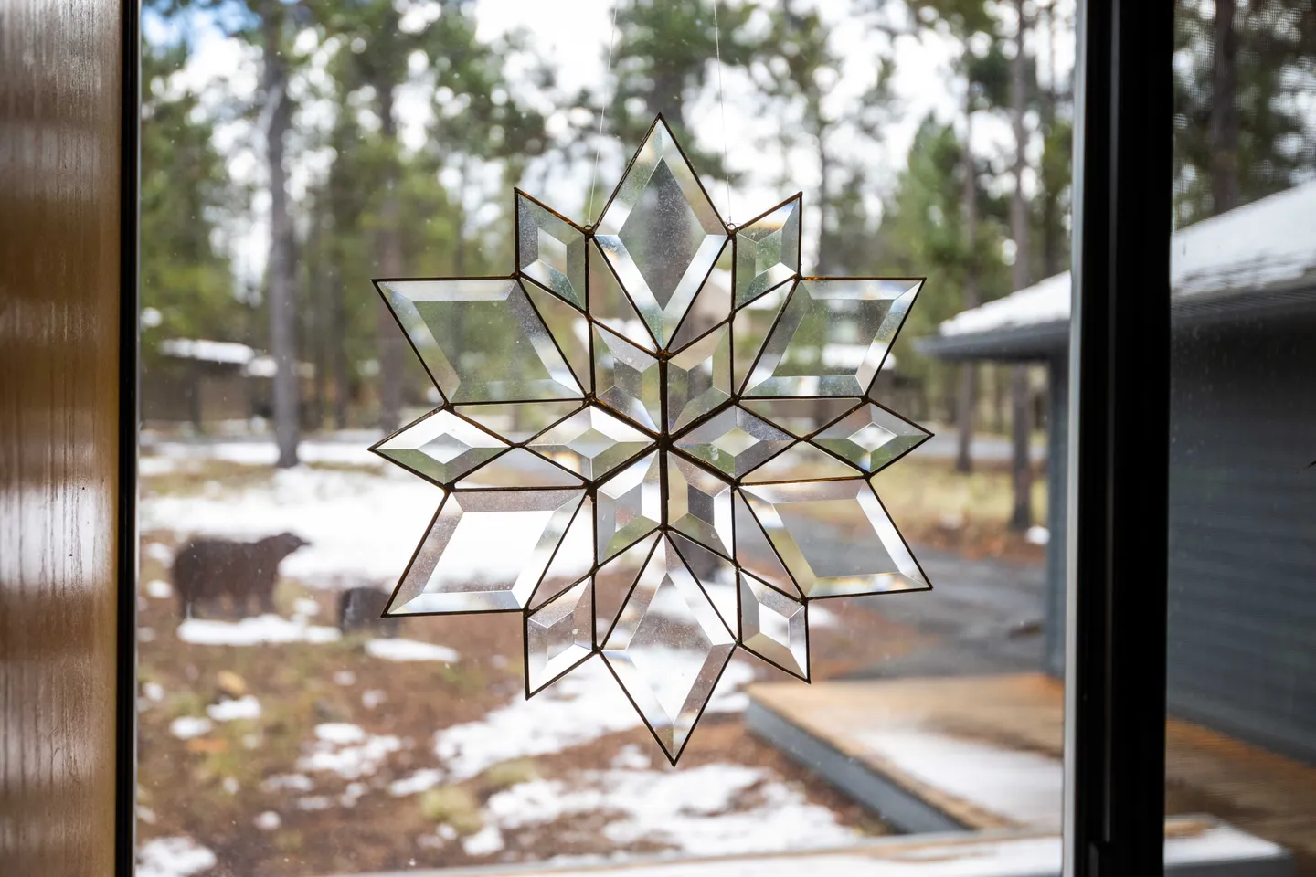 A beveled glass star suncatcher hanging in front of a window overlooking pine trees and snow.