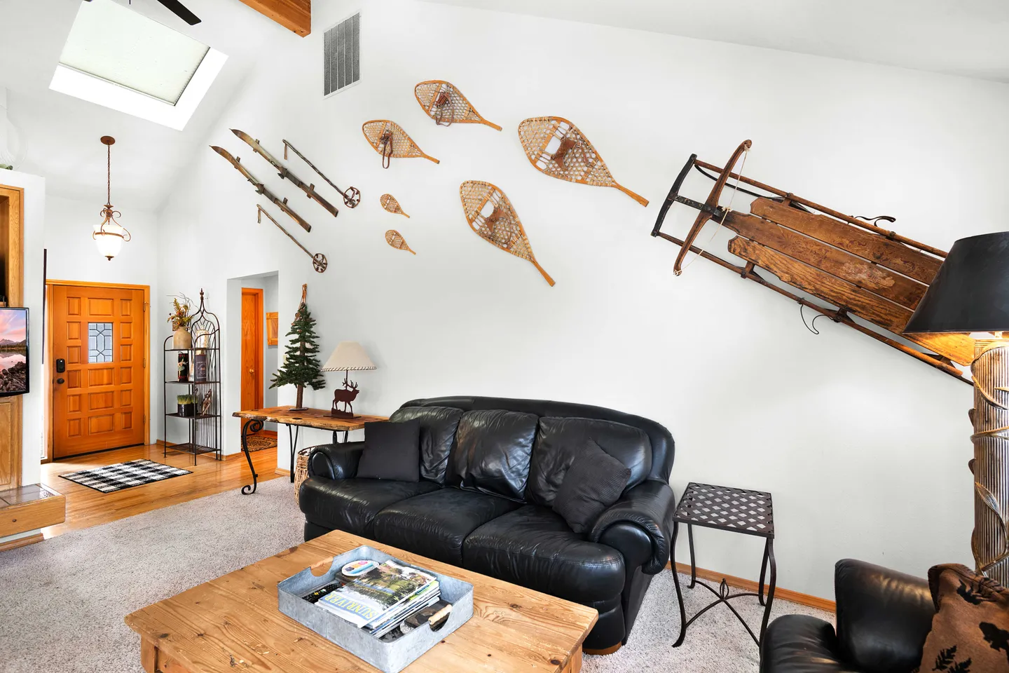 Living room with vaulted ceiling featuring winter-themed wall decor including vintage skis, snowshoes, and a wooden sled above a black leather sofa.