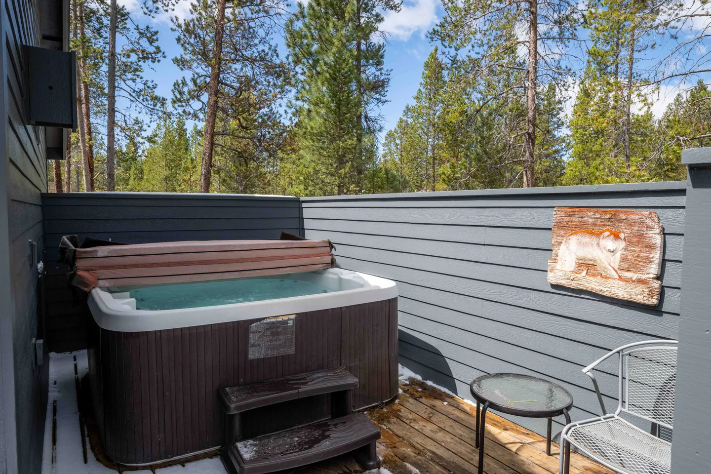 Outdoor hot tub on a wooden deck enclosed by grey privacy fencing with pine trees in the background.